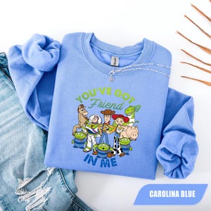 May include: Carolina blue crew neck sweatshirt featuring a graphic of Toy Story characters with the text "YOU'VE GOT A FRIEND IN ME". The sweatshirt is laid on top of a pair of blue jeans.