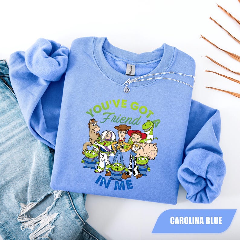 May include: Carolina blue crew neck sweatshirt featuring a graphic of Toy Story characters with the text "YOU'VE GOT A FRIEND IN ME". The sweatshirt is laid on top of a pair of blue jeans.