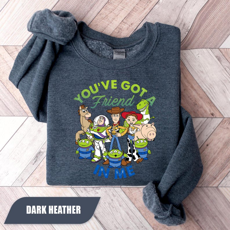 May include: Dark heather gray sweatshirt featuring a colorful graphic of characters from the movie Toy Story. The text "YOU'VE GOT A FRIEND IN ME" is printed above and below the characters. The sweatshirt is folded on a wooden surface.