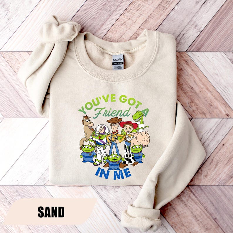 May include: Sand-colored sweatshirt featuring a graphic of characters from the movie Toy Story. The text "YOU'VE GOT A FRIEND IN ME" is printed in green and blue. The sweatshirt has a crew neck and long sleeves.