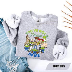 May include: Sport grey sweatshirt featuring a colorful graphic of Toy Story characters with the text "YOU'VE GOT A FRIEND IN ME". The sweatshirt has a crew neck and long sleeves. The sweatshirt is displayed on a white surface with blue jeans.