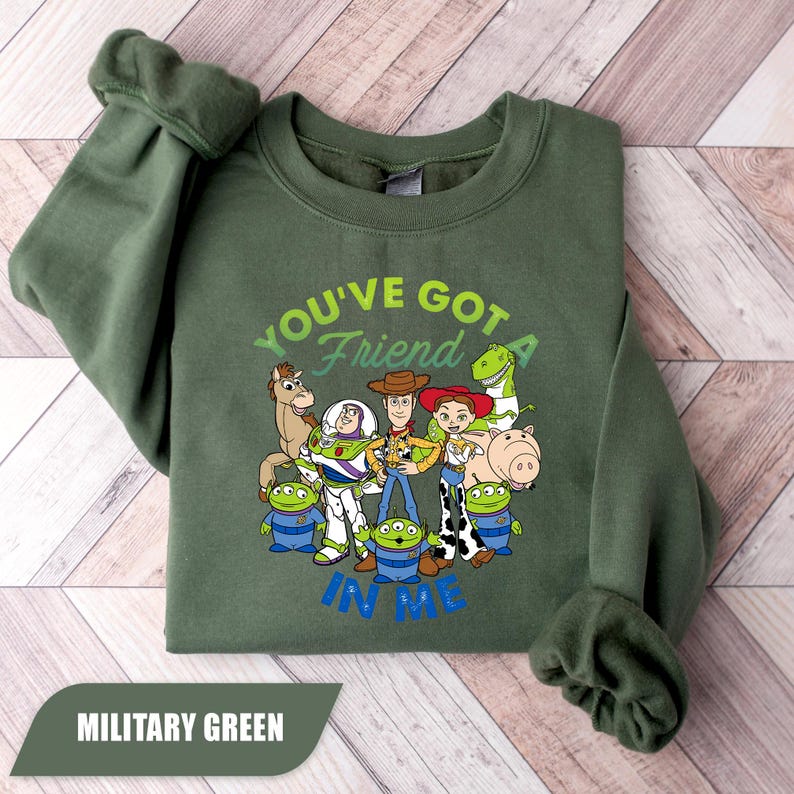 May include: Military green sweatshirt featuring a graphic of Toy Story characters with the text "YOU'VE GOT A FRIEND IN ME" in green and blue. The sweatshirt is a crew neck style.