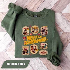 May include: Military green sweatshirt with a graphic design featuring various cartoon characters and the text "NEEEEEW ACHIEVEMENT!". The design includes a cat, a dog, a goat, and other illustrations. The sweatshirt is a solid color.