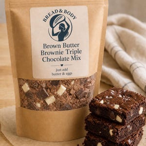 Peut inclure: Un sachet en papier kraft de préparation pour brownies au beurre noisette et triple chocolat Bread & Body, avec une fenêtre transparente montrant les ingrédients secs et des pépites de chocolat. Trois brownies au chocolat sont empilés à côté.