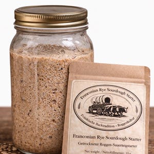 Könnte beinhalten: Ein Glas mit goldenem Deckel, gefüllt mit einem sprudelnden, braunen Sauerteig. Daneben befindet sich ein kleines Papierpäckchen mit der Aufschrift "Franconian Rye Sourdough Starter". Der Text "Made in Germany" steht oben.