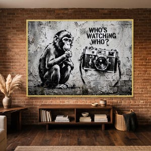 May include: A rectangular wall art print featuring a black and white stencil-style illustration of a seated chimpanzee looking at a vintage camera. The text WHO'S WATCHING WHO? is printed in black above the camera against a weathered brick wall background.