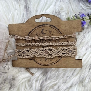 May include: Three beige macrame bracelets featuring small, round, light brown beads. The bracelets are displayed on a rectangular wooden card with a laser-engraved moon and star design. The macrame work consists of intricate knotted patterns.