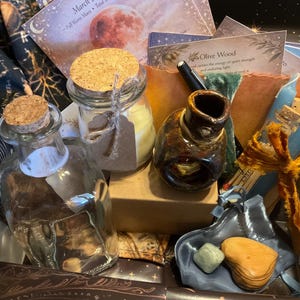 May include: A collection of spiritual items in a box, including glass bottles with cork stoppers, a ceramic inkwell, candles, and cards. The items are arranged on a patterned fabric, with a focus on natural elements and earthy tones.