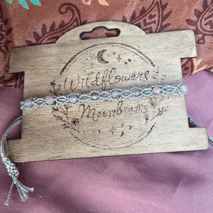 May include: A light gray macrame bracelet featuring small, round, pale pink beads woven into the cord. The bracelet is displayed on a rectangular wooden card with a laser-engraved circular design and the text Wildflowers and Moonbeams.