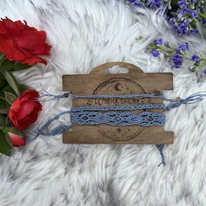 May include: Three light blue macrame bracelets wrapped around a rectangular wooden display card. The card features a laser-engraved moon, stars, and the text Wildflowers. The bracelets include intricate knotted patterns and small beads.