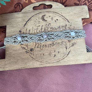 May include: A light gray macrame bracelet featuring three round, pale, iridescent beads woven into a complex knotted pattern. The bracelet is displayed on a rectangular wooden card with the text Wildflowers and Moonbeams burned into it.