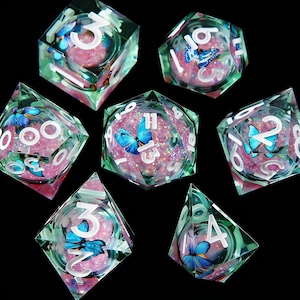 Pode incluir: Conjunto de sete dados poliédricos com tema de borboleta. Cada dado é transparente com um tom verde, preenchido com glitter rosa e apresenta uma borboleta azul e números brancos. Perfeito para jogos de tabuleiro.