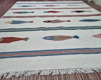 May include: A rectangular, off-white rug with a repeating pattern of colorful fish. The fish are in various colors, including blue, red, and brown. The rug has blue and red stripes and a fringed edge.