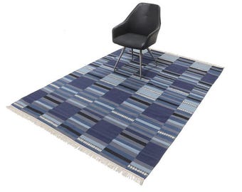 Op de afbeelding: Een rechthoekig blauw tapijt met een geometrisch patroon van horizontale strepen in verschillende tinten blauw, grijs en zwart. Het tapijt heeft een gefranjerde rand en een zwarte stoel met een leren zitting.