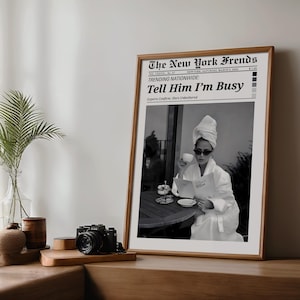 Puede incluir: Una impresión en blanco y negro enmarcada de una página de periódico titulada "The New York Frends" con el titular "Tell Him I'm Busy". La imagen dentro del marco muestra a una persona con una bata blanca y gafas de sol, sosteniendo una taza y leyendo.