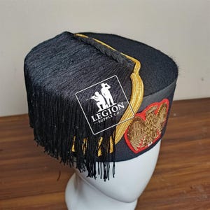 Op de afbeelding: Zwarte militaire hoed met gouden afwerking en zwarte franjes. De hoed heeft een rood en goud geborduurd embleem en een label met de tekst "LEGION SUPPLY CO."