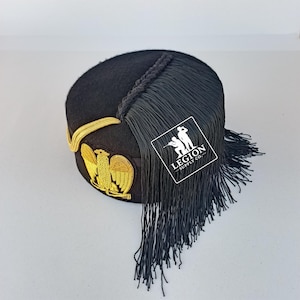 Puede incluir: Sombrero negro con emblema de águila bordado en oro y ribetes. El sombrero presenta una larga borla negra y un detalle trenzado en la parte superior. El sombrero tiene una corona redonda y una parte superior plana.