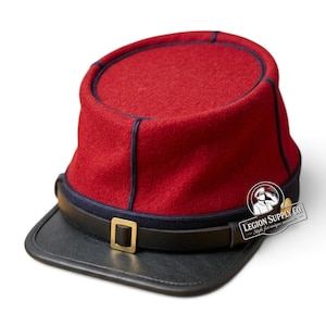 Op de afbeelding: Een rode wollen kepi-pet met donkerblauwe biezen, een zwarte lederen klep en een zwarte lederen kinband die is vastgezet met een goudkleurige metalen gesp. Een cirkelvormig logo met de tekst LEGION SUPPLY CO. Style for unique moments is zichtbaar.