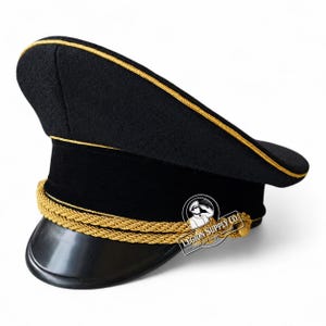 Puede incluir: Una gorra de plato negra con una corona alta y ribetes dorados en el borde superior. La gorra incluye una banda negra, una visera negra y un cordón trenzado dorado de doble capa en la parte delantera. Se ve un logotipo con el texto Legion Supply Co.