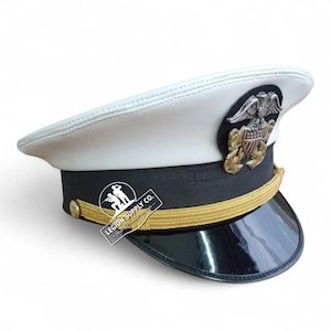 Könnte beinhalten: Eine weiße Marineoffiziersmütze mit schwarzem Band und glänzendem schwarzem Schirm. Die Mütze hat ein goldfarbenes Emblem und Zierleisten sowie ein kleines Etikett mit der Aufschrift "LEGION SUPPLY CO."