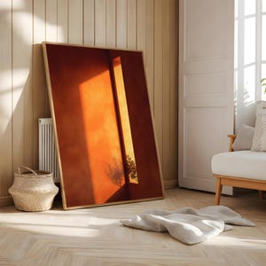 May include: A large framed art print featuring an abstract image of a sunlit interior. The artwork leans against a light wood-paneled wall. A woven basket and a white radiator are visible. The room has a neutral color palette.