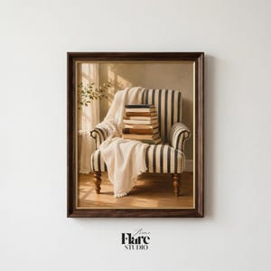 May include: A framed print of a cozy reading nook. A striped armchair holds a stack of books and a draped blanket. The artwork is in a dark brown frame, with the "Frame Flare Studio" logo at the bottom.