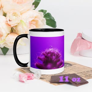 Puede incluir: Taza de cerámica blanca con asa y borde negros, con un primer plano de un cactus morado con una gota de agua. La taza tiene una capacidad de 325 ml. El fondo incluye flores y un trozo de chocolate.
