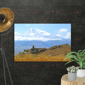 Puede incluir: Una impresión de paisaje con una montaña nevada bajo un cielo azul con nubes. Una pequeña iglesia con tejado rojo se encuentra en una colina en primer plano, con hierba seca delante. Una bandera ondea en la cima de la colina.