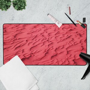 May include: A rectangular desk mat with a red, textured surface resembling sand dunes. The mat is surrounded by beauty products, including lipstick, a tube of cream, and a black eyeliner pencil. A white towel and a black hair dryer are also present.