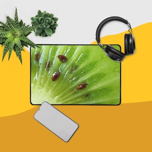 May include: A rectangular mousepad featuring a close-up of a kiwi fruit slice, with a bright green color and visible brown seeds. Black headphones and a smartphone are placed on the side. Two small potted succulents are also visible.