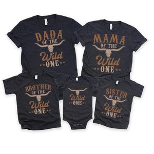 May include: A set of five dark heather grey shirts and a bodysuit featuring a tan bull skull graphic and text. The shirts read DADA OF THE WILD ONE, MAMA OF THE WILD ONE, BROTHER OF THE WILD ONE, SISTER OF THE WILD ONE, and the bodysuit says WILD ONE.