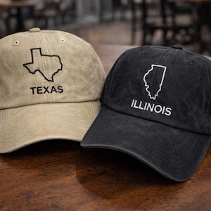 May include: Two baseball caps, one tan and one black, are displayed on a wooden surface. The tan cap features a Texas outline and the word "TEXAS." The black cap has an Illinois outline and the word "ILLINOIS."