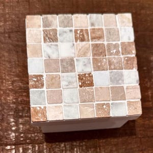 Pode incluir: Uma caixa decorativa quadrada, com mosaicos e base branca. Os mosaicos são pequenos quadrados em tons de bege, castanho e branco. A caixa está sobre uma superfície de madeira.