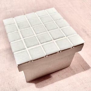 Pode incluir: Uma pequena caixa retangular com uma parte superior de mosaico. As telhas quadradas são brancas sujas, dispostas em um padrão de grade. As laterais da caixa são de uma cor clara e neutra, com uma sombra em uma superfície rosa.
