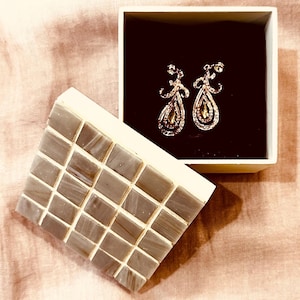 Pode incluir: Um par de brincos pendurados em forma de gota, com um centro dourado e detalhes brilhantes, apresentados numa caixa branca com interior preto. Uma base de azulejo cinzento está ao lado.