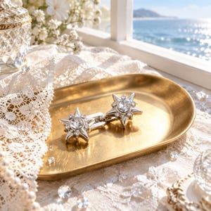 May include: A pair of silver-toned star-shaped cufflinks featuring a central clear round rhinestone surrounded by smaller inset crystals. The star design has pointed rays and a polished metallic finish, presented on a smooth gold-toned oval tray.