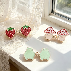 May include: Three pairs of decorative pins: strawberries, mushrooms, and frogs. The strawberry pins are red with green leaves. The mushroom pins have red and white caps. The frog pins are light green. All pins are connected by a gold-colored bar.