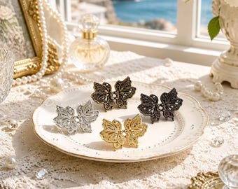 May include: Four butterfly-shaped hair clips in silver, gold, bronze, and dark gunmetal tones. Each clip features intricate, openwork filigree wings with a symmetrical design, arranged on a white ceramic plate with a scalloped edge and gold trim.