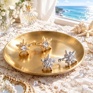 May include: Two pairs of star-shaped earrings, one gold-toned and one silver-toned, each featuring a central clear crystal surrounded by smaller accent stones. The earrings are displayed on a round, brushed gold-toned metal tray.