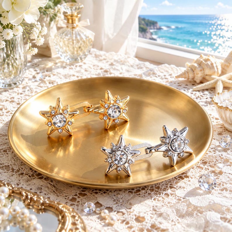 May include: Two pairs of star-shaped earrings, one gold-toned and one silver-toned, each featuring a central clear crystal surrounded by smaller accent stones. The earrings are displayed on a round, brushed gold-toned metal tray.