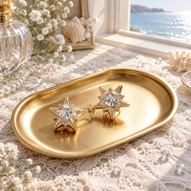 May include: A pair of gold-toned star-shaped earrings featuring a central clear crystal surrounded by smaller inset crystals. The earrings are resting on a smooth, oval-shaped gold metal tray with a polished finish and a raised edge.