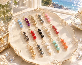 May include: A collection of fifteen metal safety pins arranged on a decorative scalloped tray. Each pin features three round beads in various colors including blue, purple, gold, grey, pink, red, black, white, orange, and teal, accented with small rhinestones.