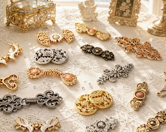 May include: A collection of ornate metal garment fasteners and decorative clasps in gold, silver, and bronze tones. These intricate pieces feature floral motifs, filigree patterns, and small inset cabochons, arranged on a white lace fabric surface.