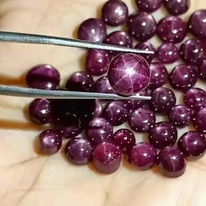 Könnte beinhalten: Eine Sammlung tiefroter Sternrubine, einige mit einem sechsstrahligen Sterneffekt. Ein Rubin wird von einer Pinzette gehalten, die seine polierte Oberfläche und sein Sternmuster zeigt. Die Edelsteine sind auf hellem Hintergrund verstreut, was ihre satte Farbe und ihren Glanz hervorhebt.