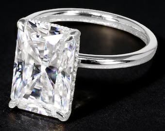 Puede incluir: Anillo de plata con un gran diamante de talla rectangular. El diamante está sujeto por cuatro garras. La banda es lisa y pulida. El anillo se muestra sobre un fondo negro.