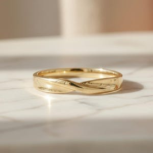 May include: A gold ring with a twisted design. The ring is made of a shiny, reflective metal and sits on a white surface. The ring's design features a continuous, interwoven pattern, creating a unique and elegant look.