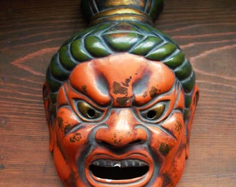 May include: An antique Japanese mask with an angry expression. The mask is predominantly orange with green and gold detailing. It has open eye holes and a wide-open mouth, suggesting a fierce character.