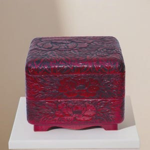 Puede incluir: Una caja cuadrada de laca roja profunda con intrincadas tallas florales de tonos oscuros en la tapa y los laterales. La caja presenta un diseño escalonado y se apoya sobre cuatro pequeñas patas integradas, mostrando un relieve detallado.