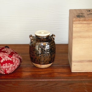 May include: A red and white patterned fabric item, a glazed ceramic jar with a lid, and a wooden box are arranged on a dark wooden surface. The jar has two handles and a light-colored base. The box has Japanese text.