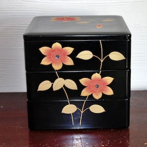 Op de afbeelding: Een zwarte, drielaagse Japanse bento-box met een glanzende afwerking. De doos is versierd met oranje en gele bloemmotieven en goudkleurige stelen en bladeren. De bloemen hebben een blauw midden.